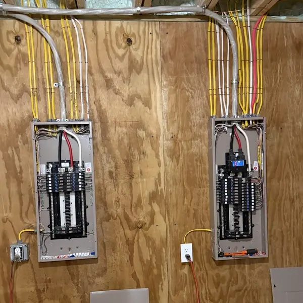 Two new breaker panels side by side on a plywood wall with the feeders and branch circuits roughed in on a new build
